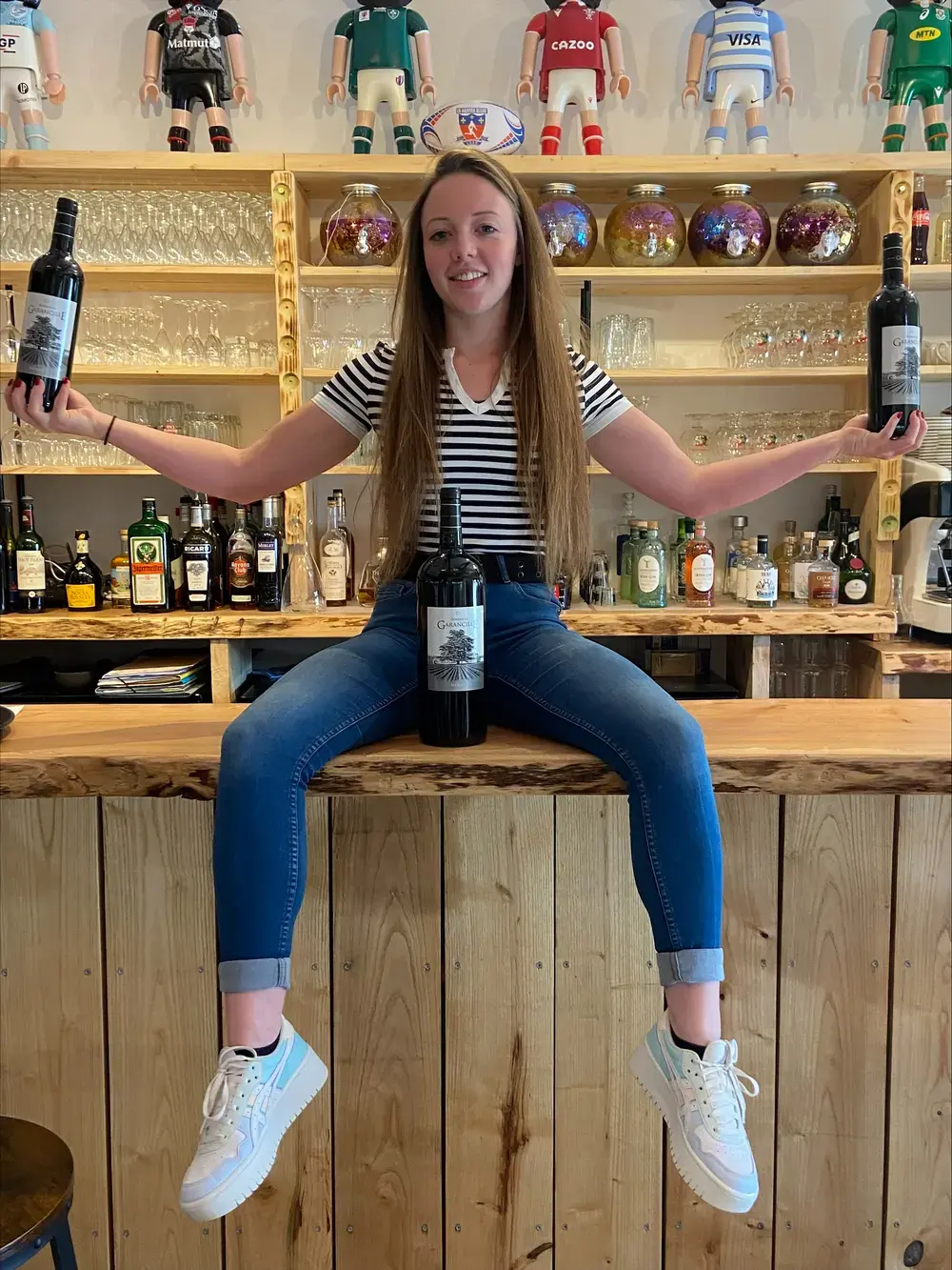 Une membre de l’équipe assise sur le bar, une bouteille de vin dans chaque main.