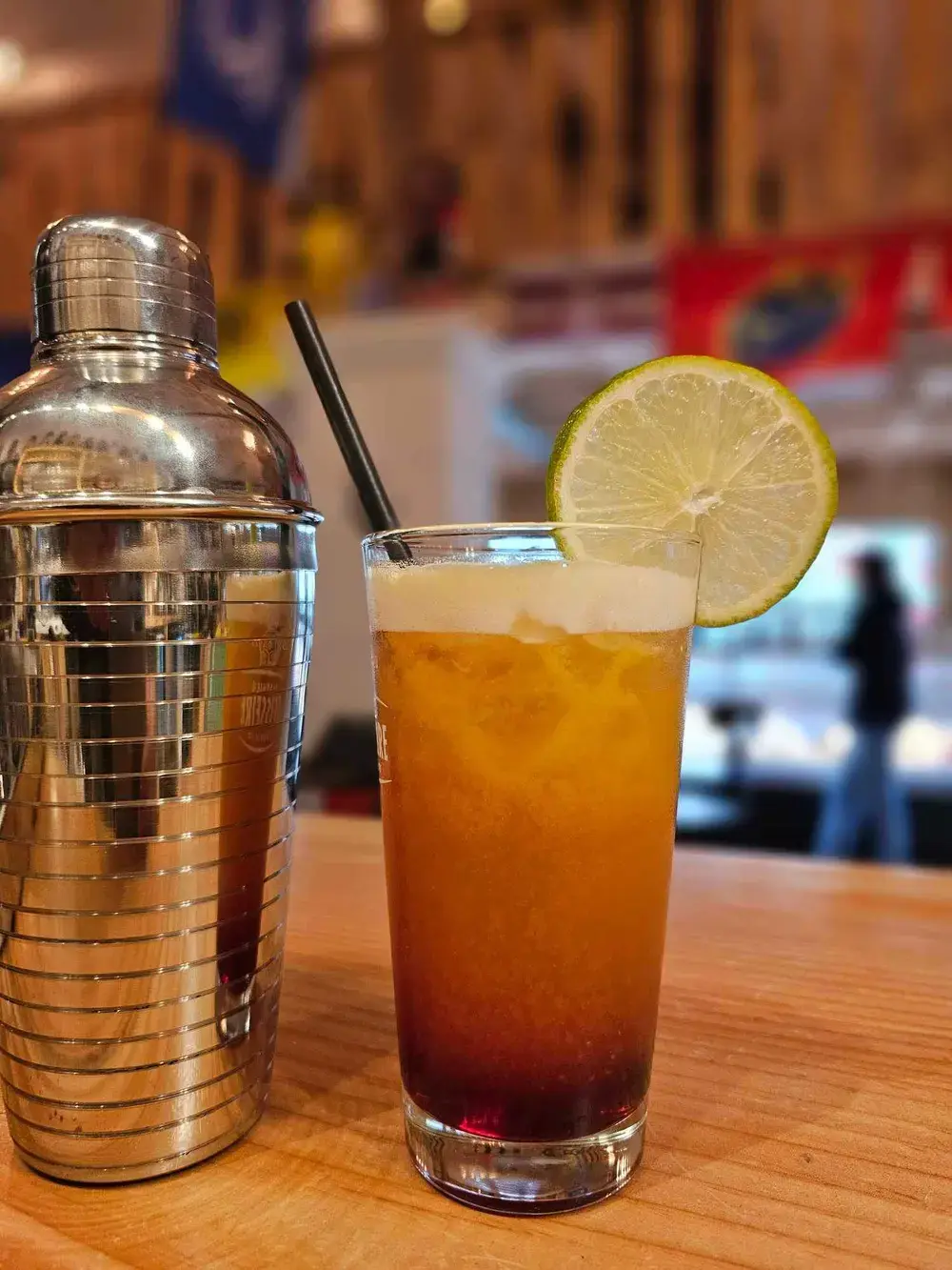 Un shaker en inox et un cocktail long drink avec sa rondelle de citron, au comptoir.
