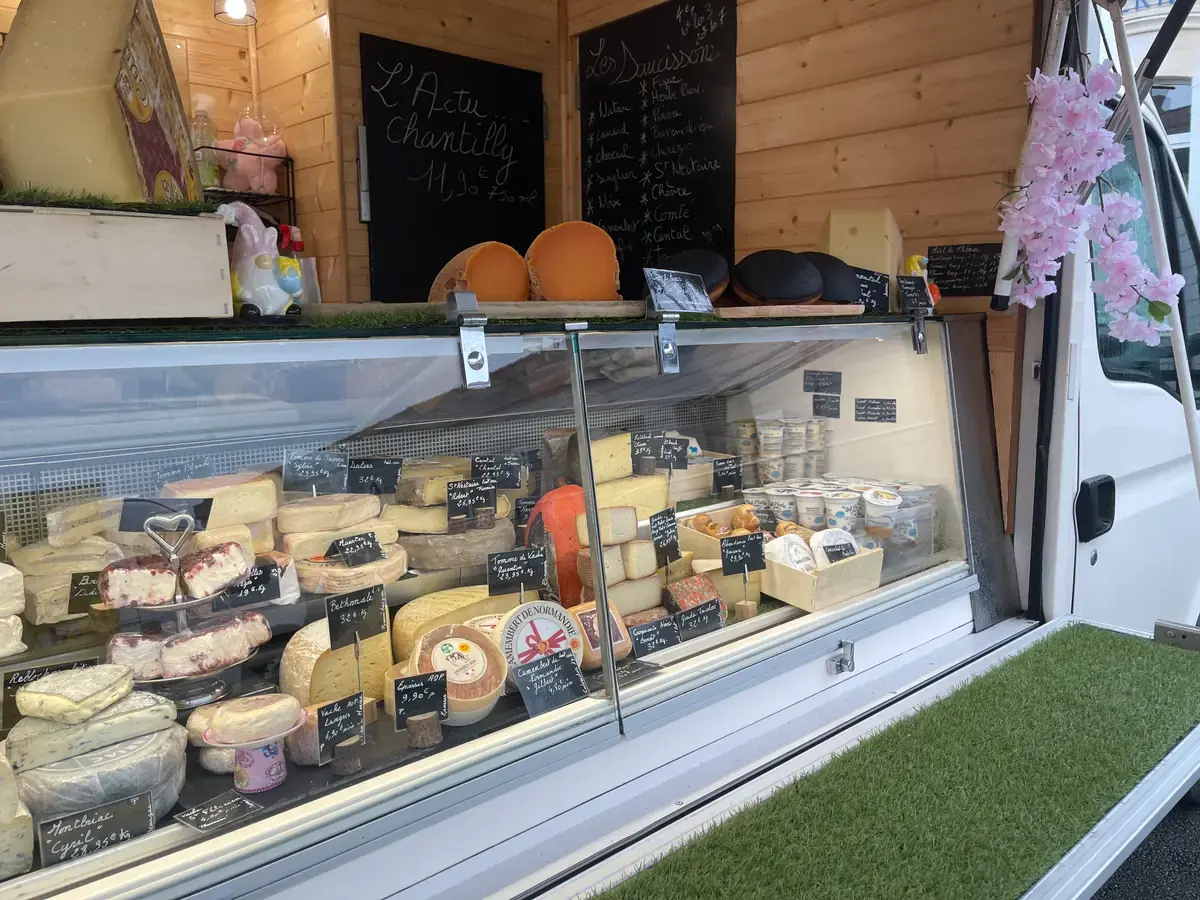 L’étal de fromages du camion vu de trois quarts, une branche de fleurs accrochée à la portière.