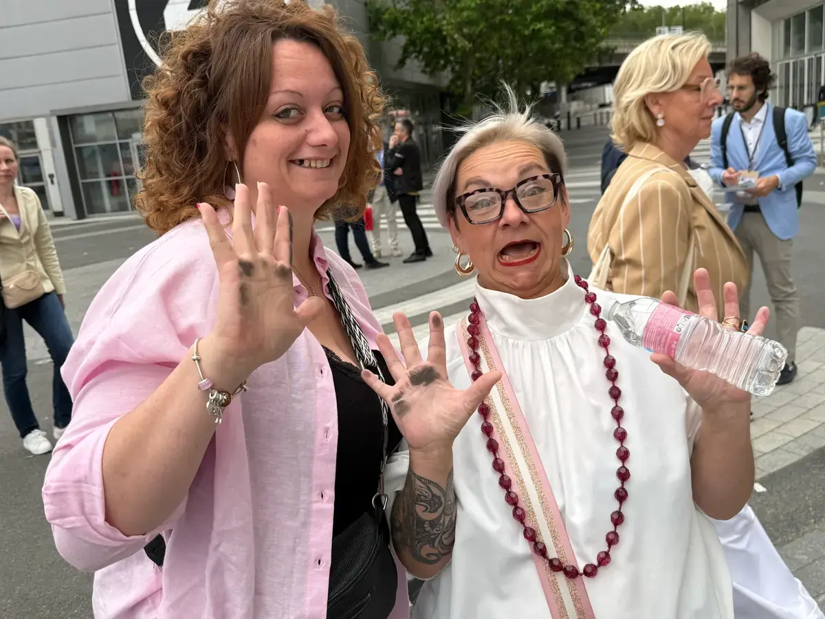 Deux femmes rient face à l’objectif, les paumes levées, sur une place de ville.