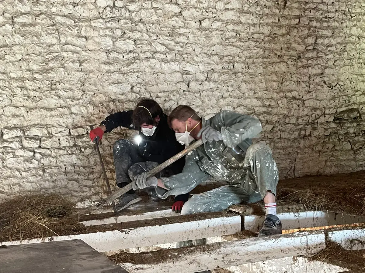 Deux compagnons masqués, à genoux devant le mur de pierre, arrachent à la barre à mine les planches et le torchis du plancher.