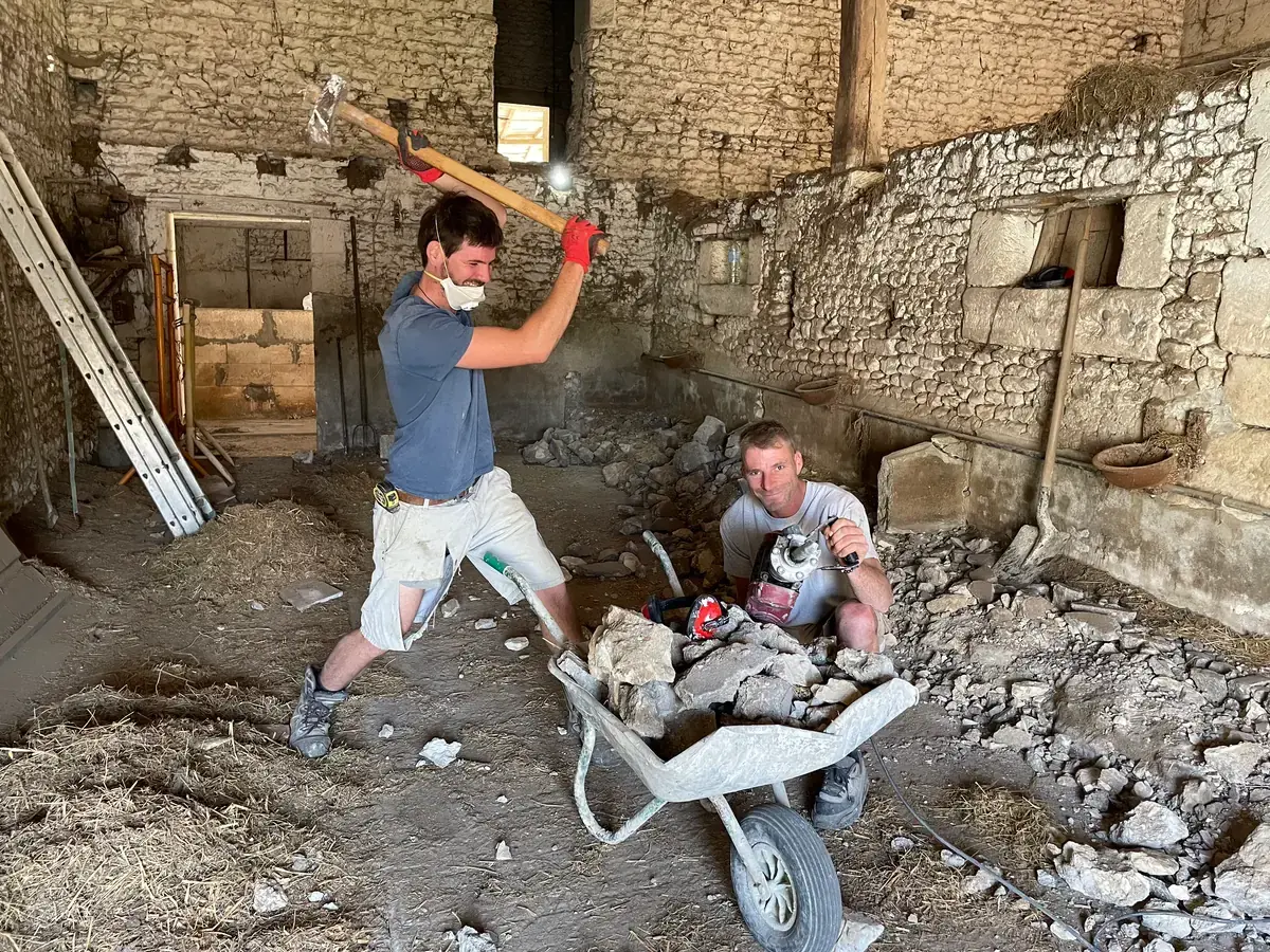 Deux hommes déblaient la grange à la pioche et à la brouette, au milieu des gravats, devant les murs de pierre mis à nu.
