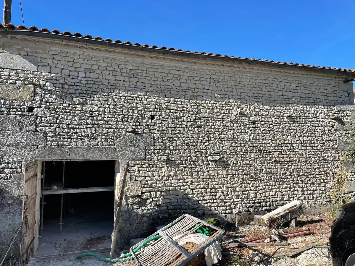 La façade en pierre de la grange sous un ciel bleu, sa grande porte ouverte sur le chantier.