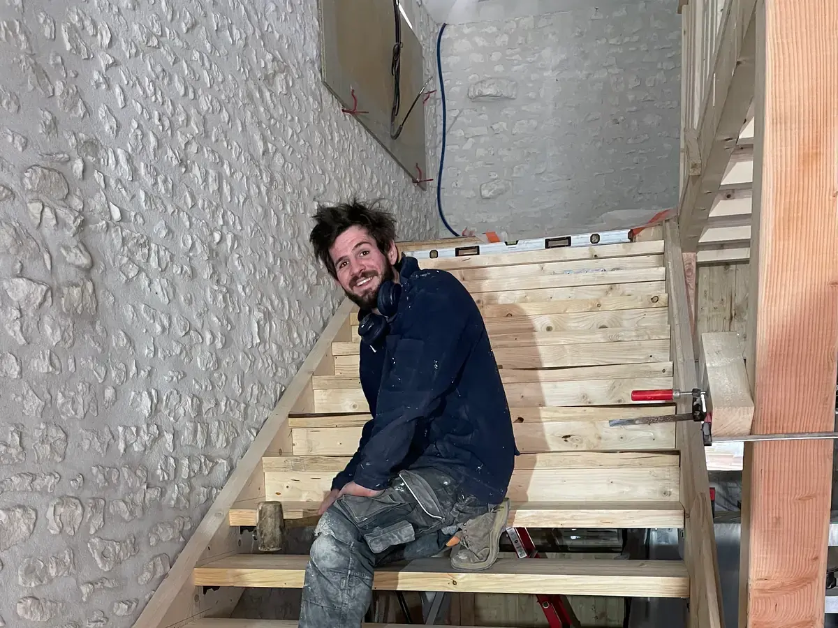 Un compagnon souriant, assis sur les marches de l’escalier de bois qu’il vient de monter contre le mur de pierre.
